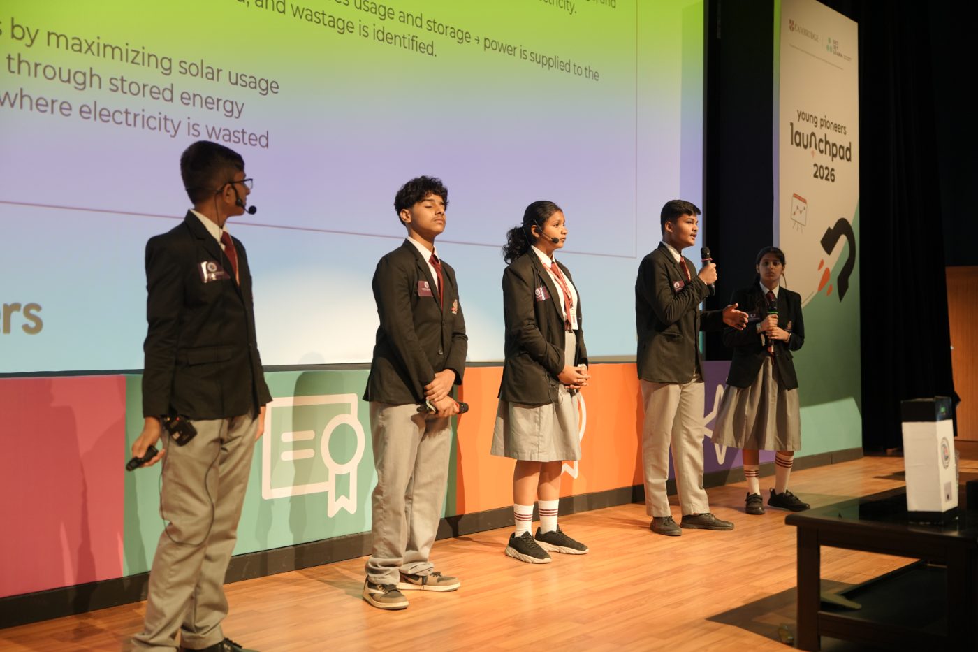 Five students on stage pitching a solar energy idea, with their slide behind them